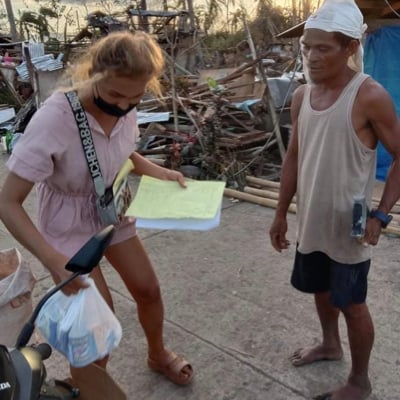 A woman wearing a mask distributes relief goods to a man in front of debris in a disaster-stricken area.